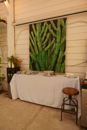 A buffet table with white tablecloth set against a wall featuring large green botanical artwork and a wooden stool.