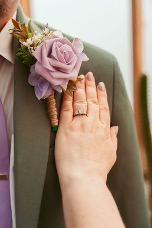 A delicate pink rose boutonniere on a green suit jacket with a hand displaying an engagement ring.