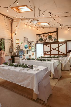 A rustic wedding reception venue with string lights, white tables, and a gallery wall of framed artwork.