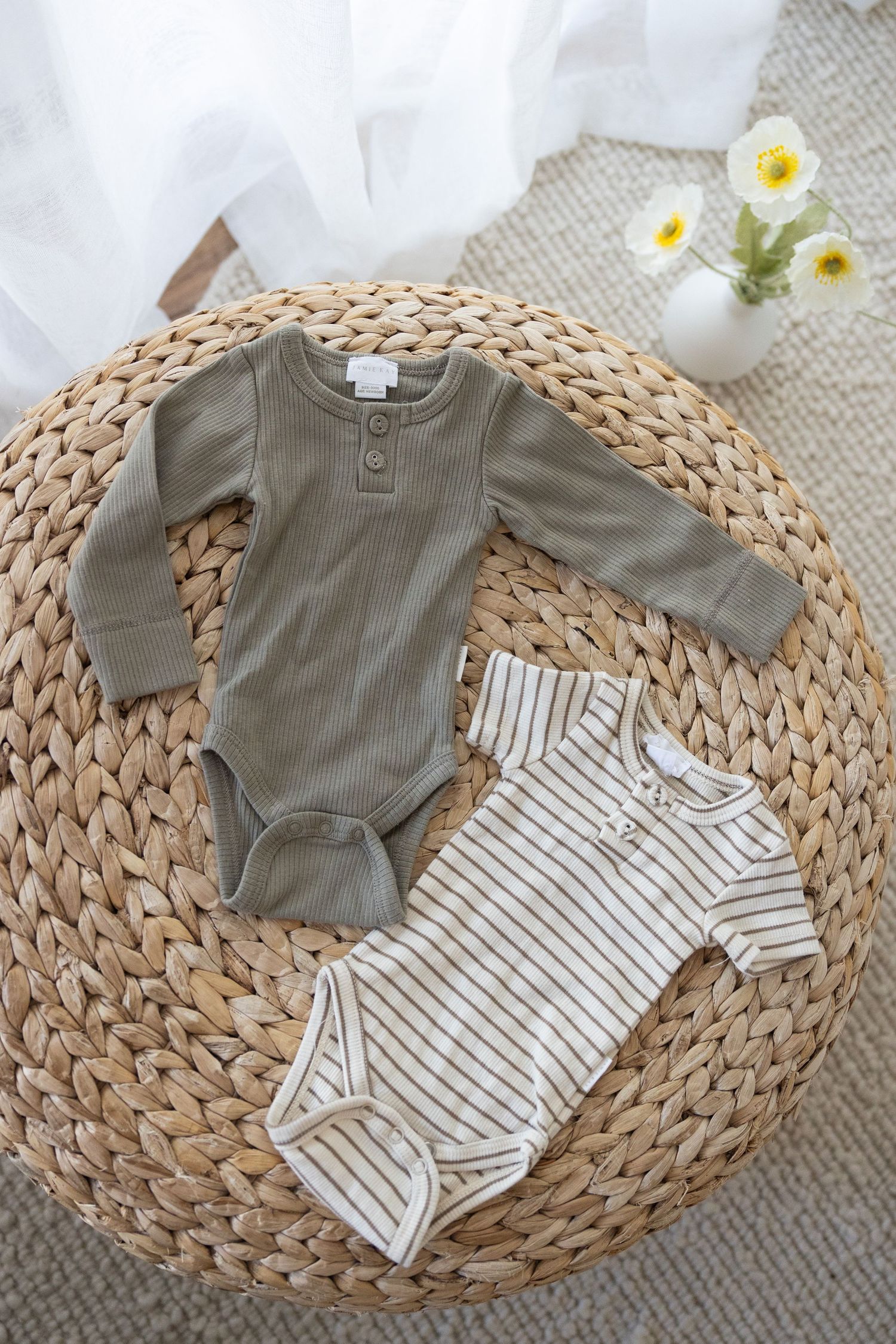 Baby bodysuits in olive green and striped patterns displayed on a woven rattan mat with small white flowers in a vase.