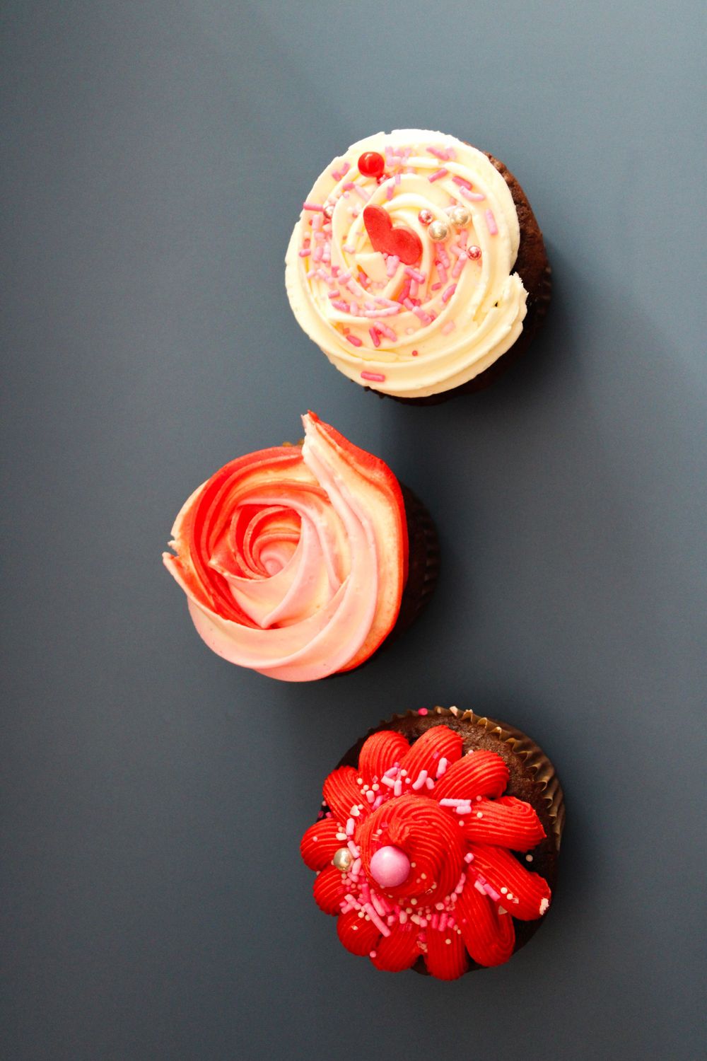 Three pink and red frosted cupcakes arranged vertically on a dark background.