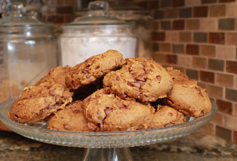 Homemade chocolate chip cookies piled on a glass cake stand against a brick wall background.