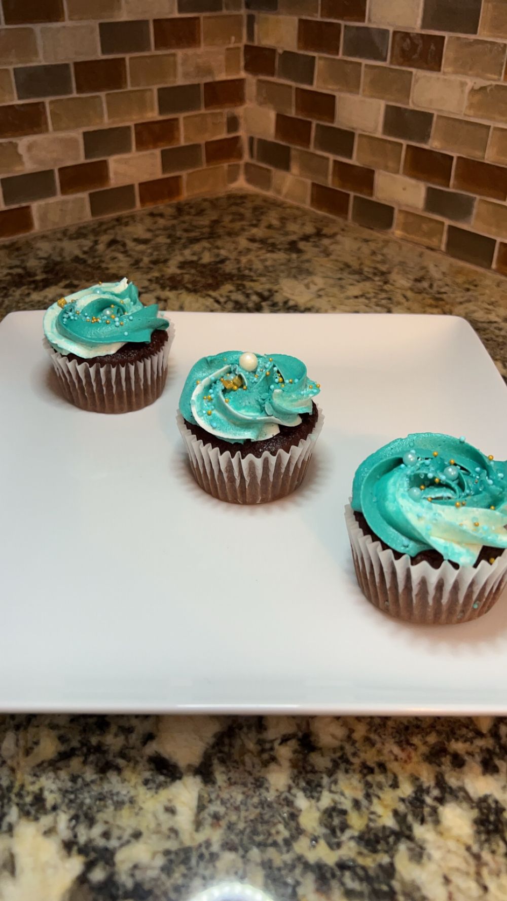 Three chocolate cupcakes with turquoise frosting and gold sprinkles on a white square plate.