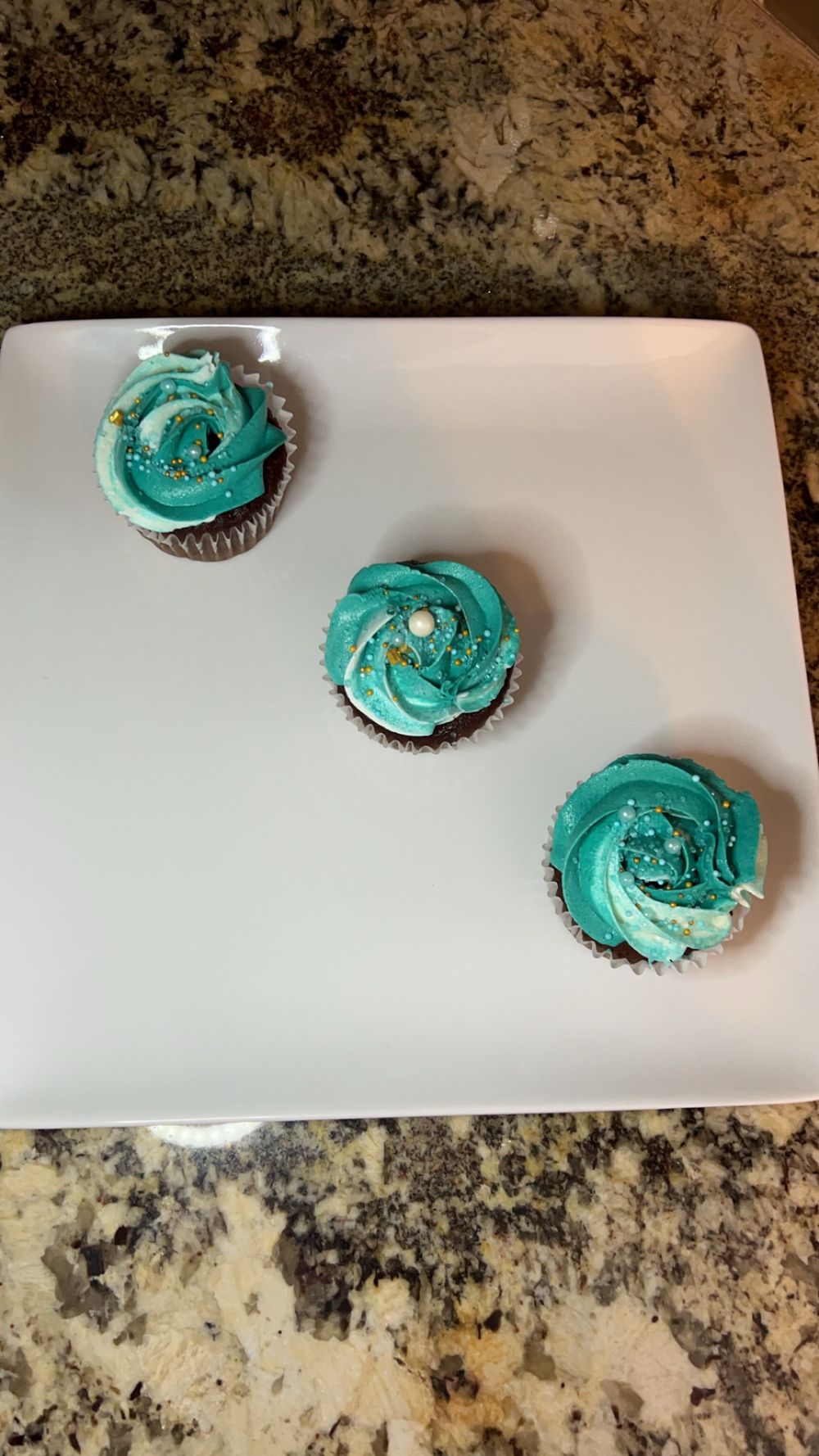 Chocolate cupcakes topped with bright turquoise frosting and gold sprinkles displayed on white ceramic plate.
