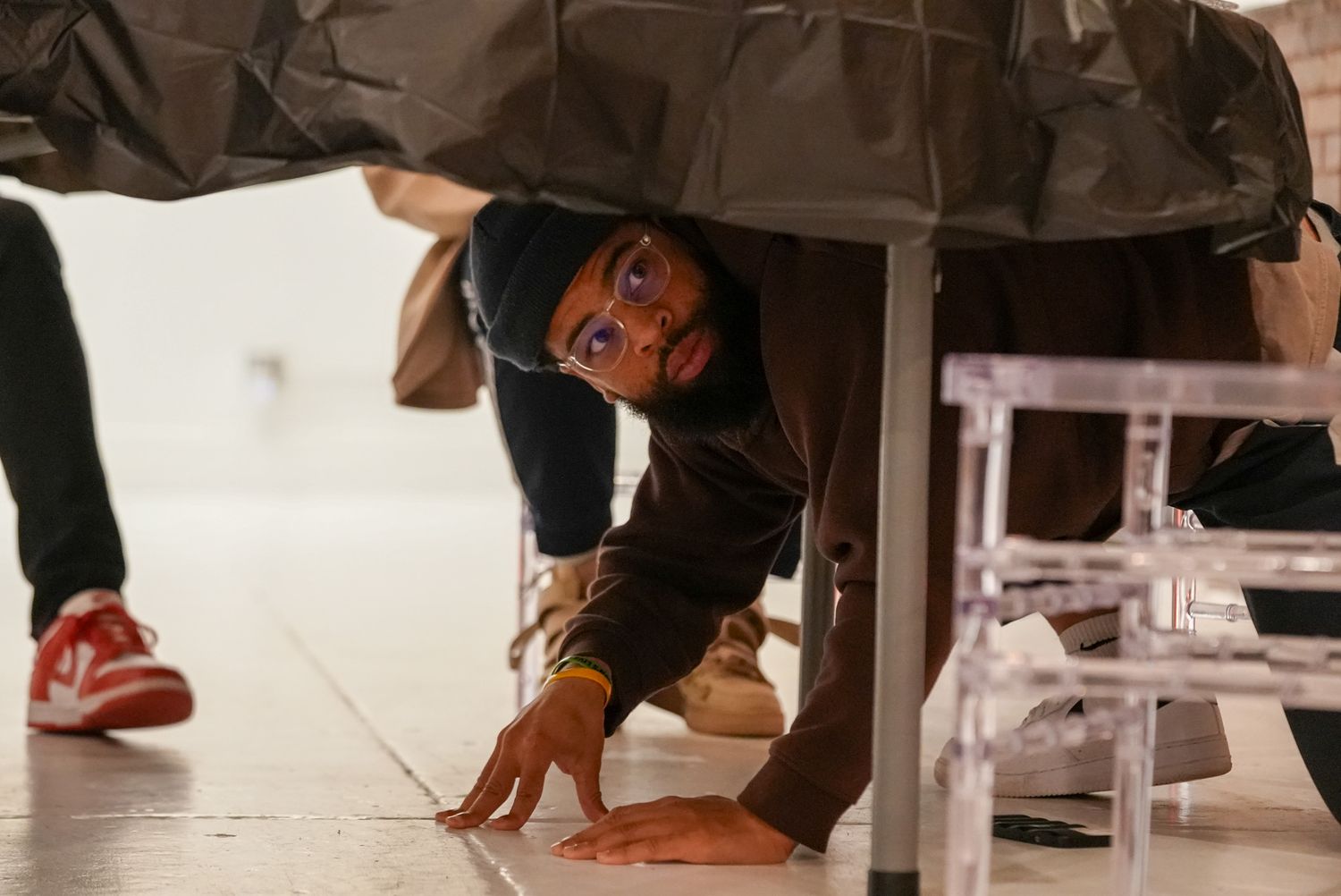 Curious child peeking under a table at an indoor event, wearing orange glasses.