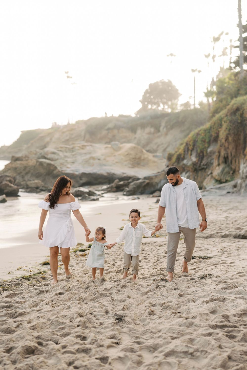 Family wearing white and beige outfits walking on a sandy beach near coastal cliffs at sunset.