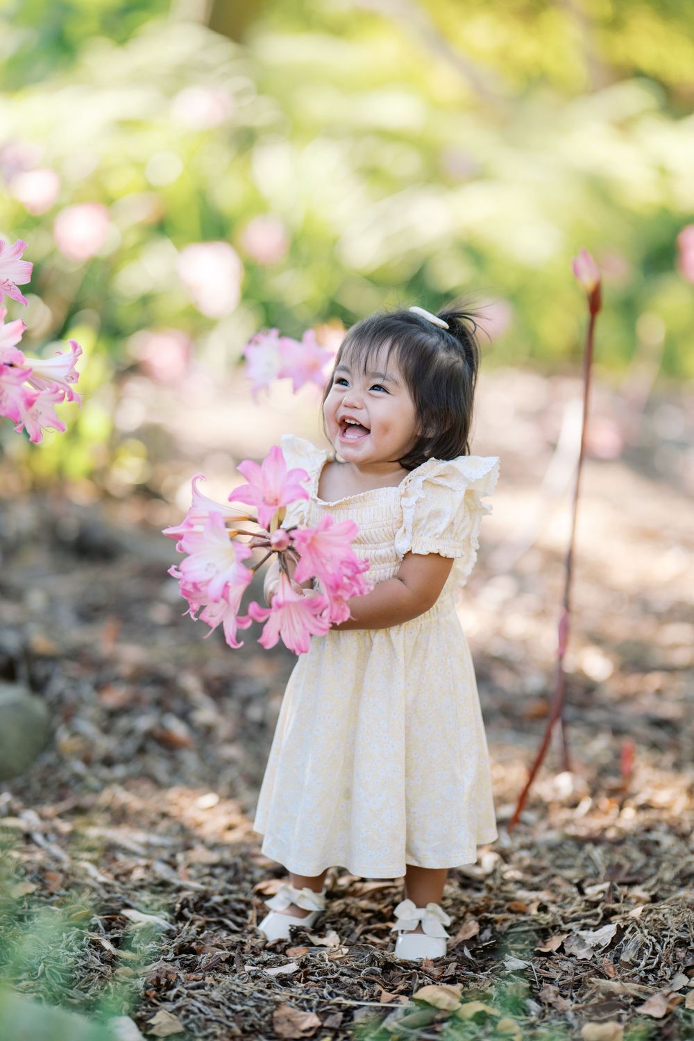 Joyful child in a white dress plays and laughs while surrounded by pink flowers in a garden setting.