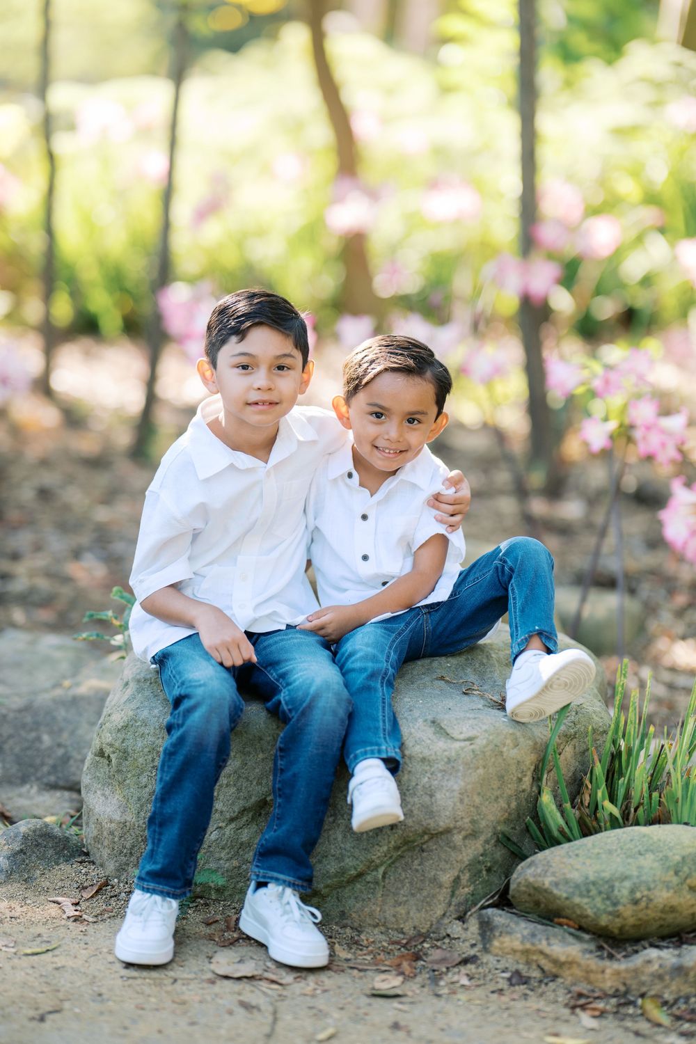 Two young siblings in white shirts and jeans sit together on natural stone steps among pink flowers in a garden setting.