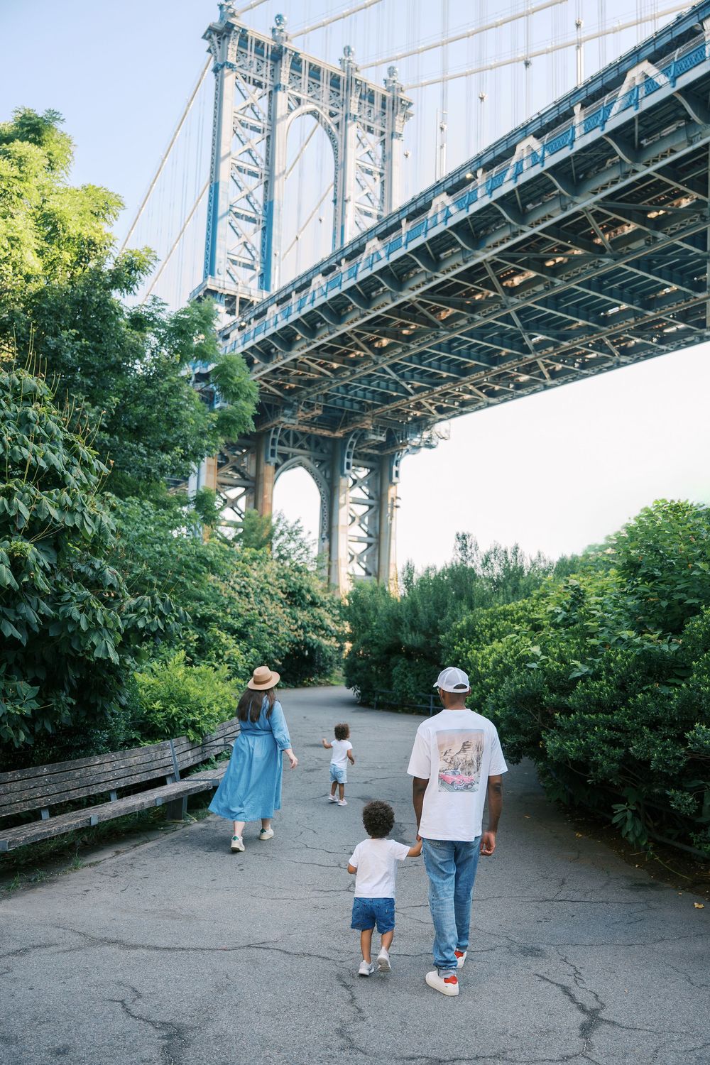 People walking on a path beneath the towering blue steel structure of a bridge on a sunny summer day.