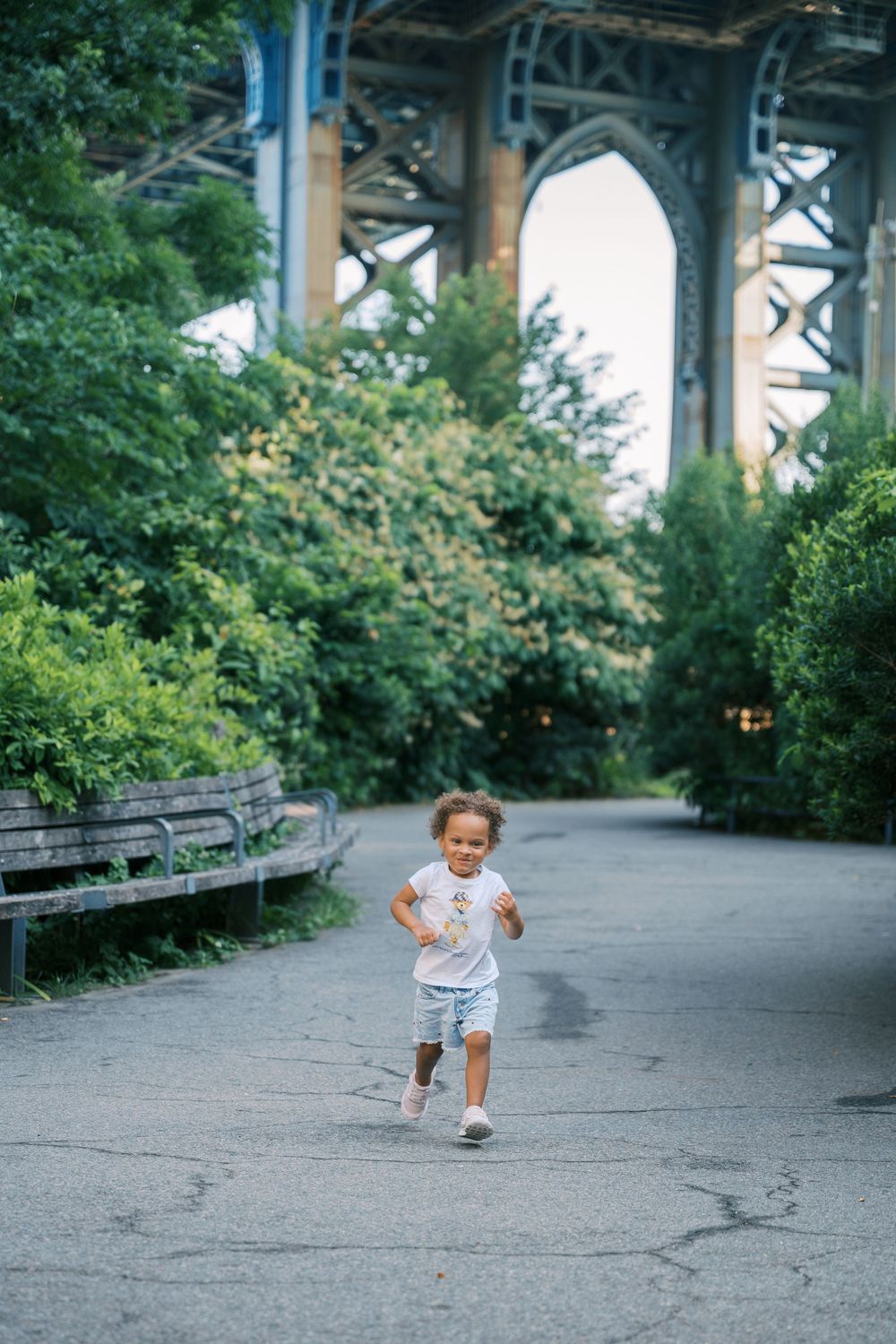 Child in white t-shirt crouches and plays in shadow of urban bridge on concrete path surrounded by greenery.