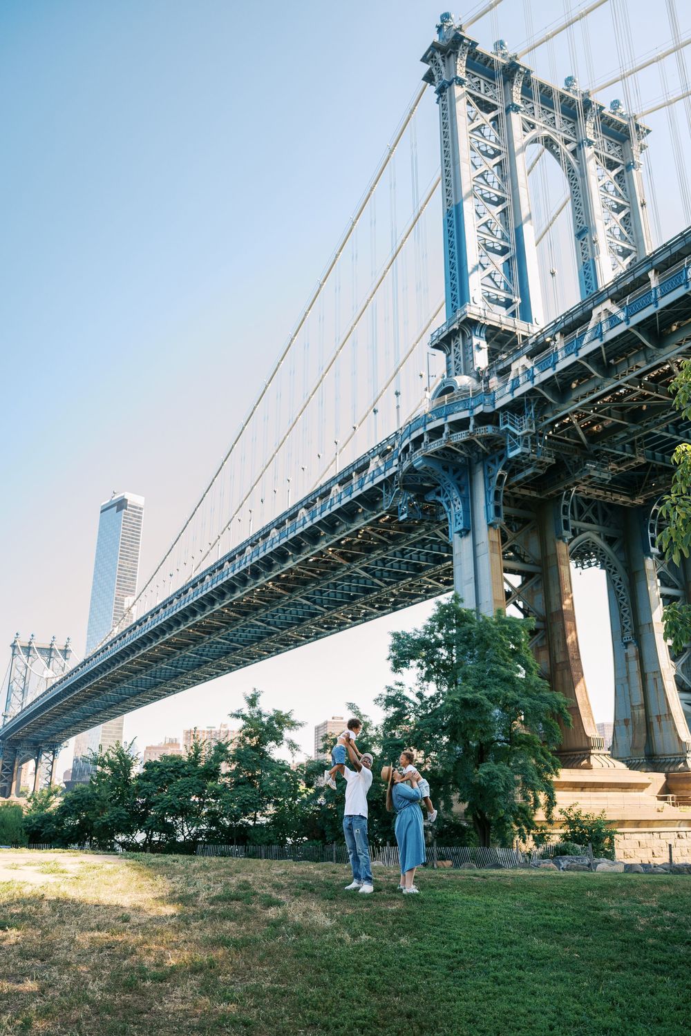 Visitors stand beneath the iconic Manhattan Bridge in Brooklyn's DUMBO neighborhood with a city skyscraper visible in background.
