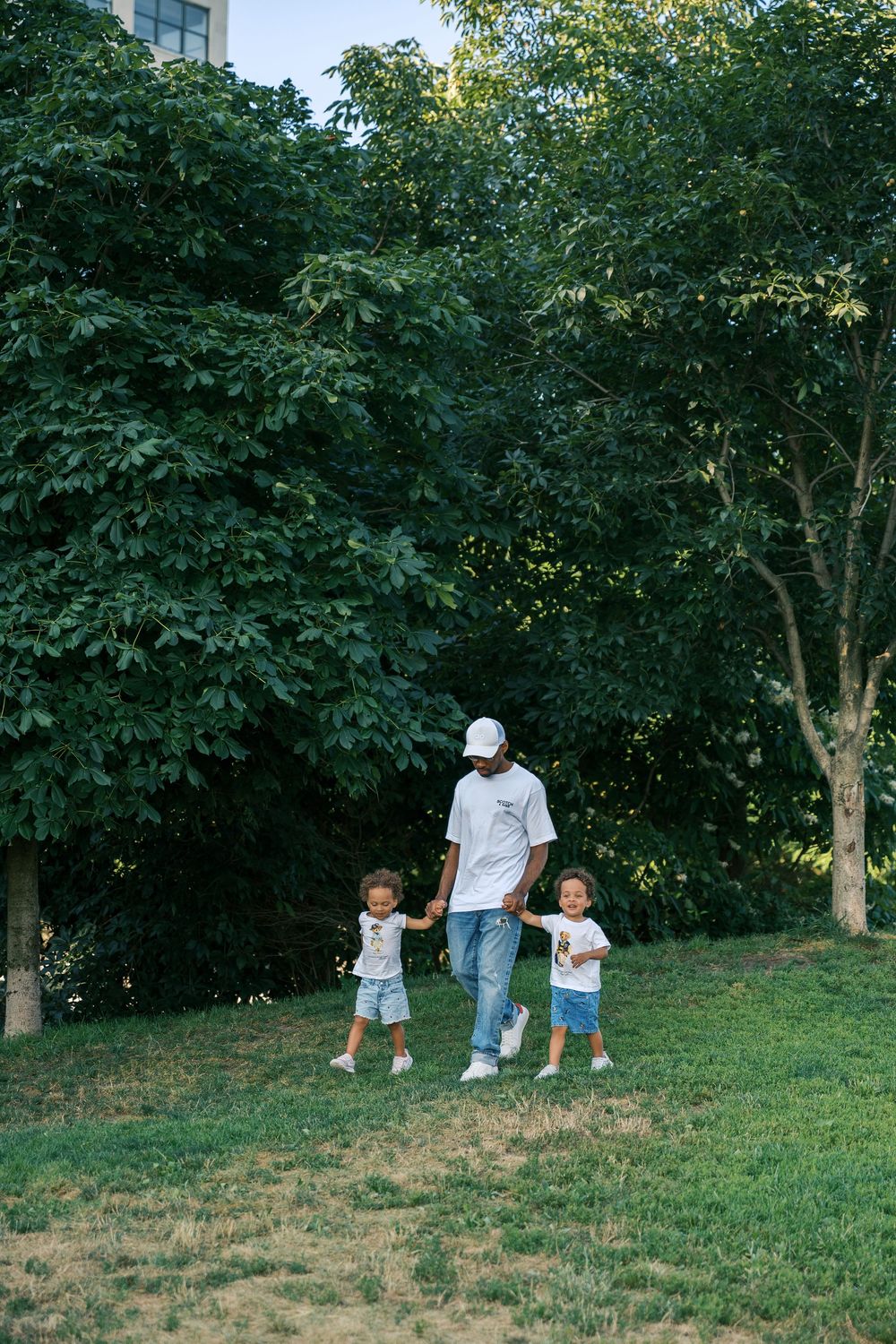 Adult and small children walk and play together on a grassy hillside surrounded by leafy green trees on a summer day.