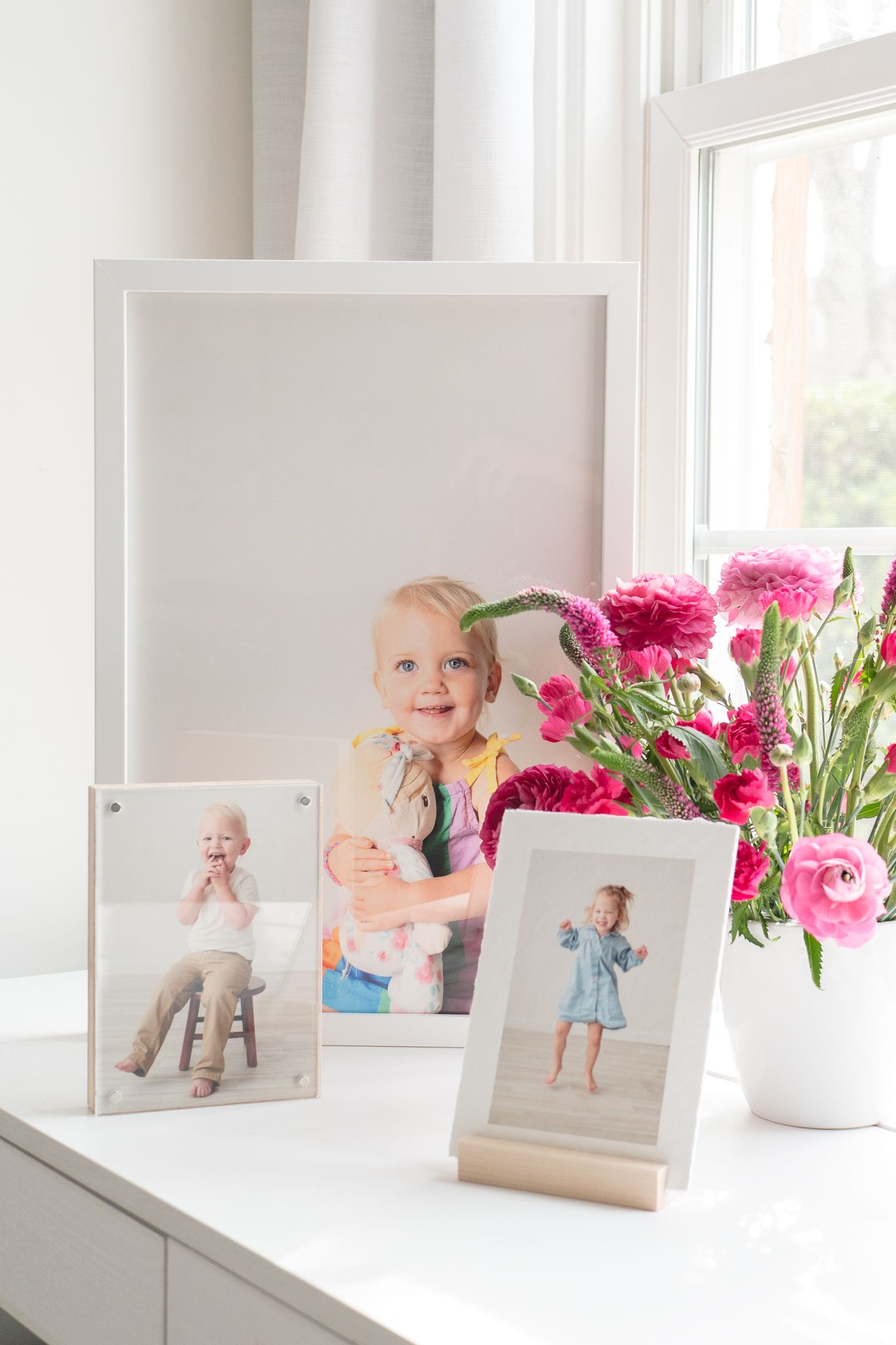 Framed family photos displayed on white windowsill with pink ranunculus flowers in white vase.