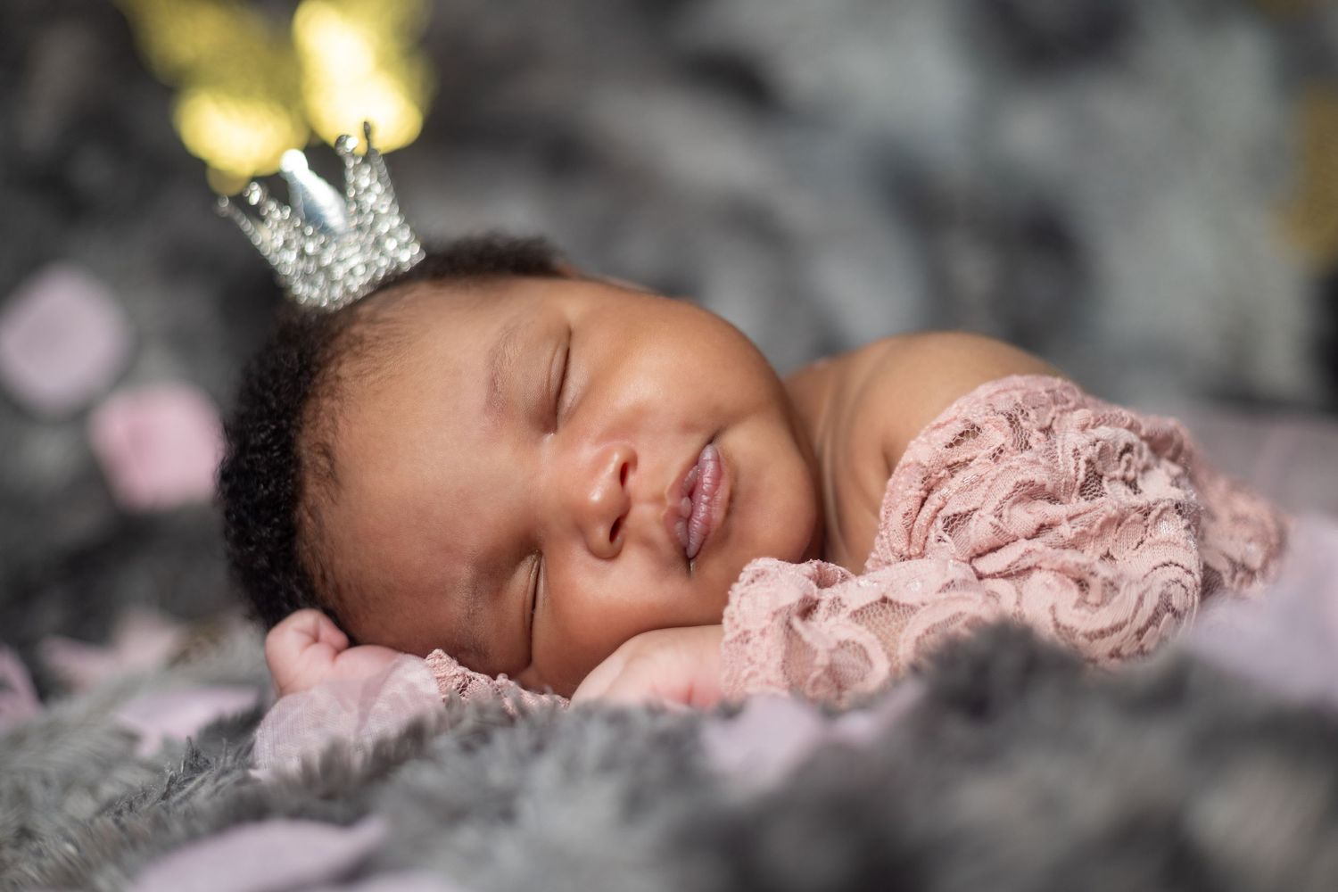 Sleeping newborn wearing tiny crown rests on soft gray blanket with bokeh lights in background.