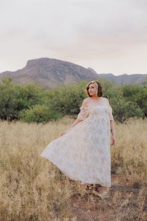 Posing in tall desert grass wearing an off-shoulder white dress with mountains behind.