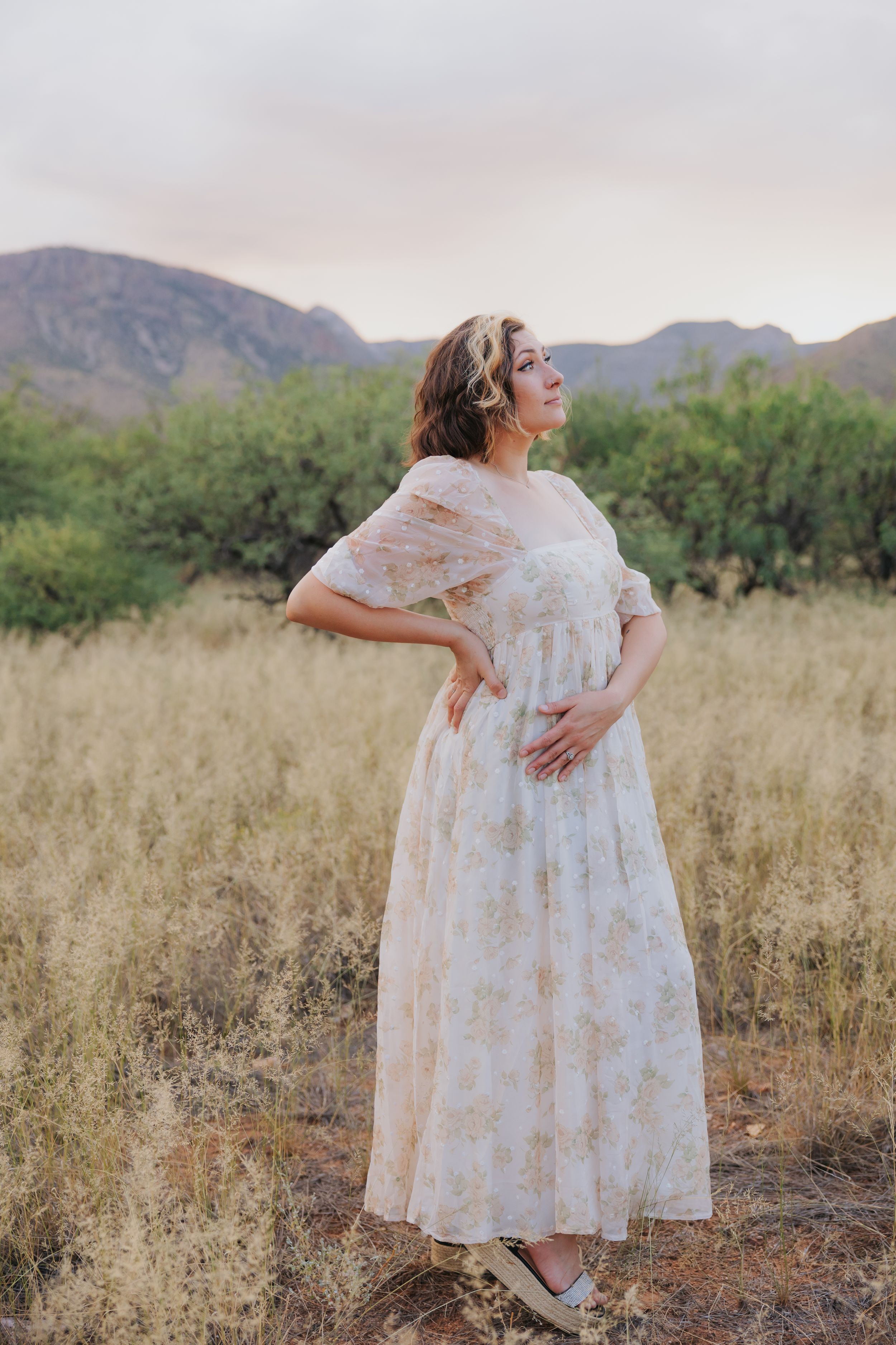 Side profile pose in a white floral dress against golden desert grass and mountains.