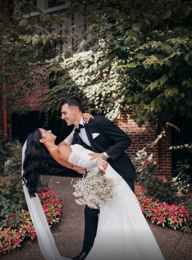 Elegant couple in wedding attire share romantic dip pose in garden setting with red brick wall and blooming flowers.
