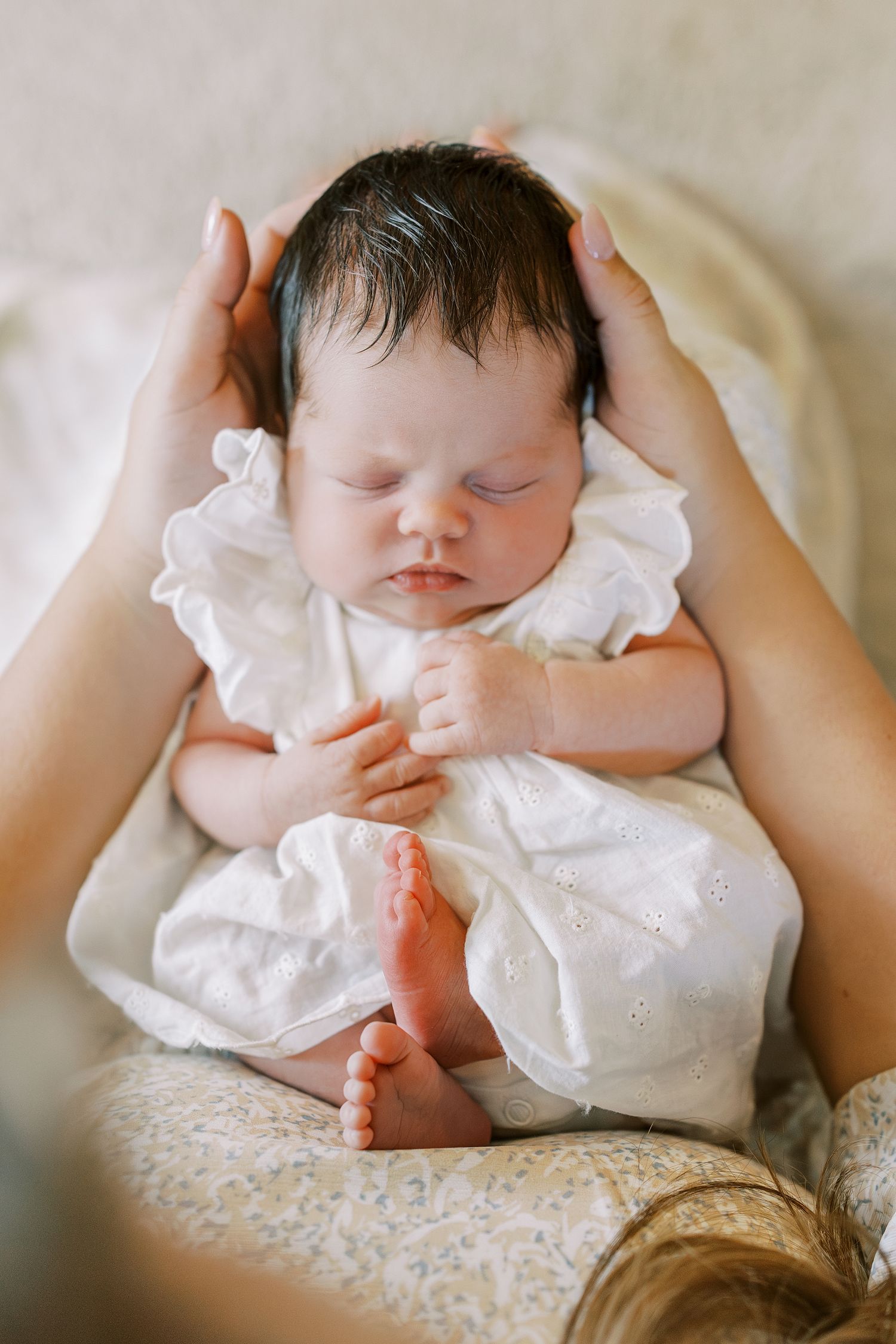 Newborn baby in white dress sleeping peacefully while being cradled in mother's hands.