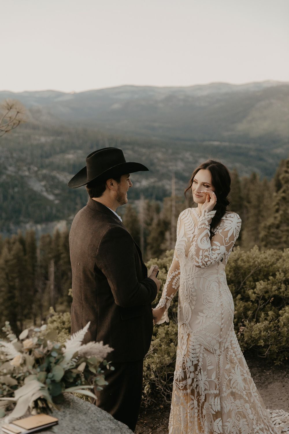 A romantic mountain wedding photoshoot featuring a couple in a lace dress and western attire against scenic forest views.