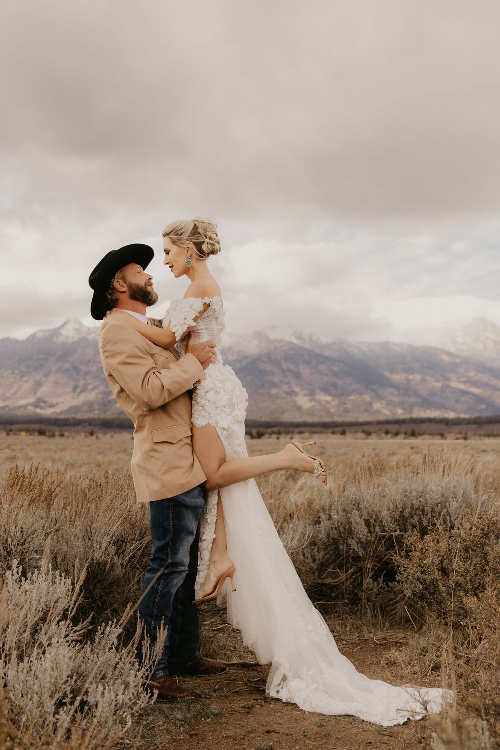 A romantic wedding couple embraces in a field with mountains in the background during an overcast day.