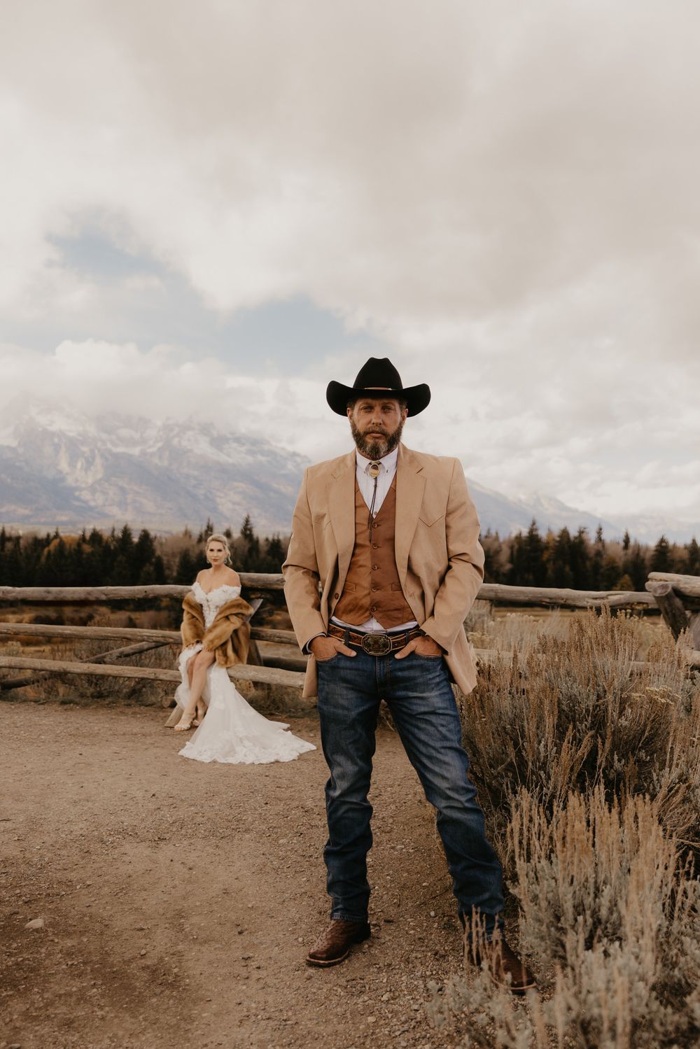A rustic western wedding photoshoot in a field with mountains and sagebrush against cloudy skies.