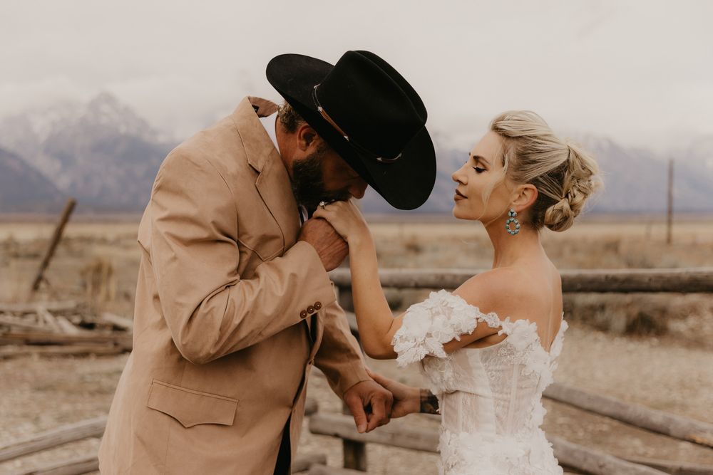 A romantic couple in western attire embraces on train tracks in a desert landscape at sunset.