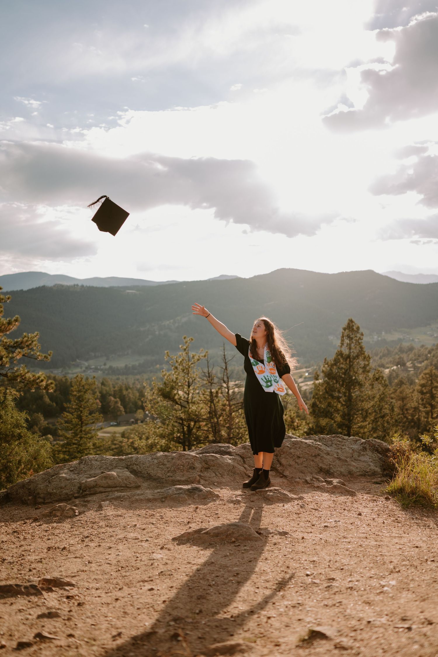 Graduate wearing a white stole tosses her black graduation cap in the air at a mountain overlook.