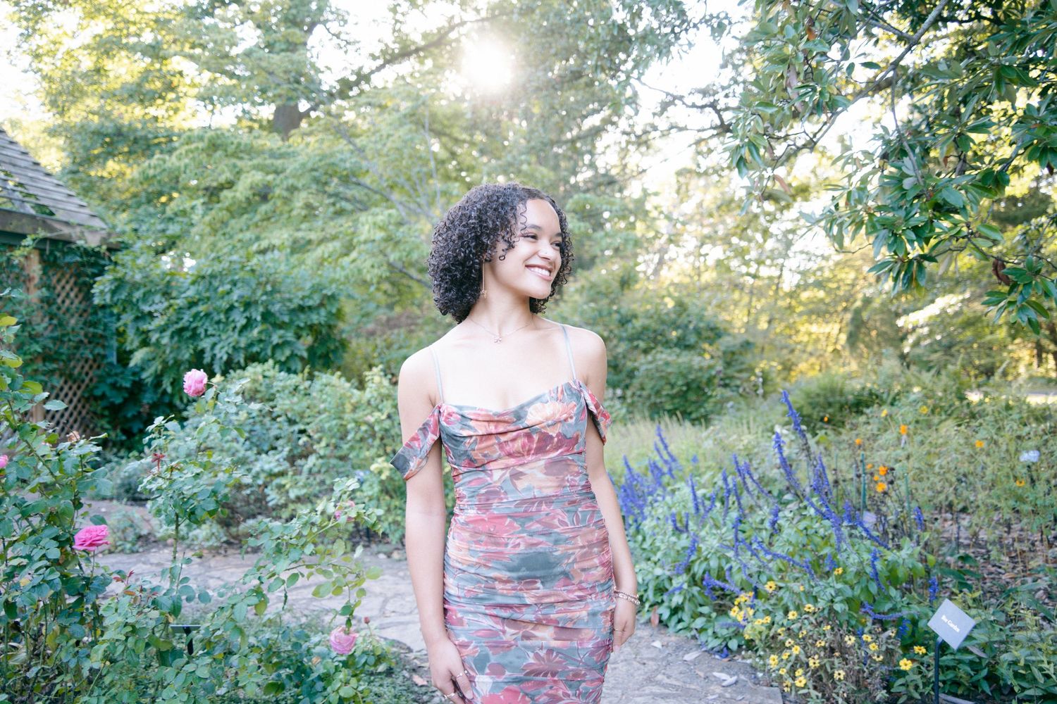 Young woman in floral off-shoulder dress smiling in lush garden with colorful flowers and greenery.