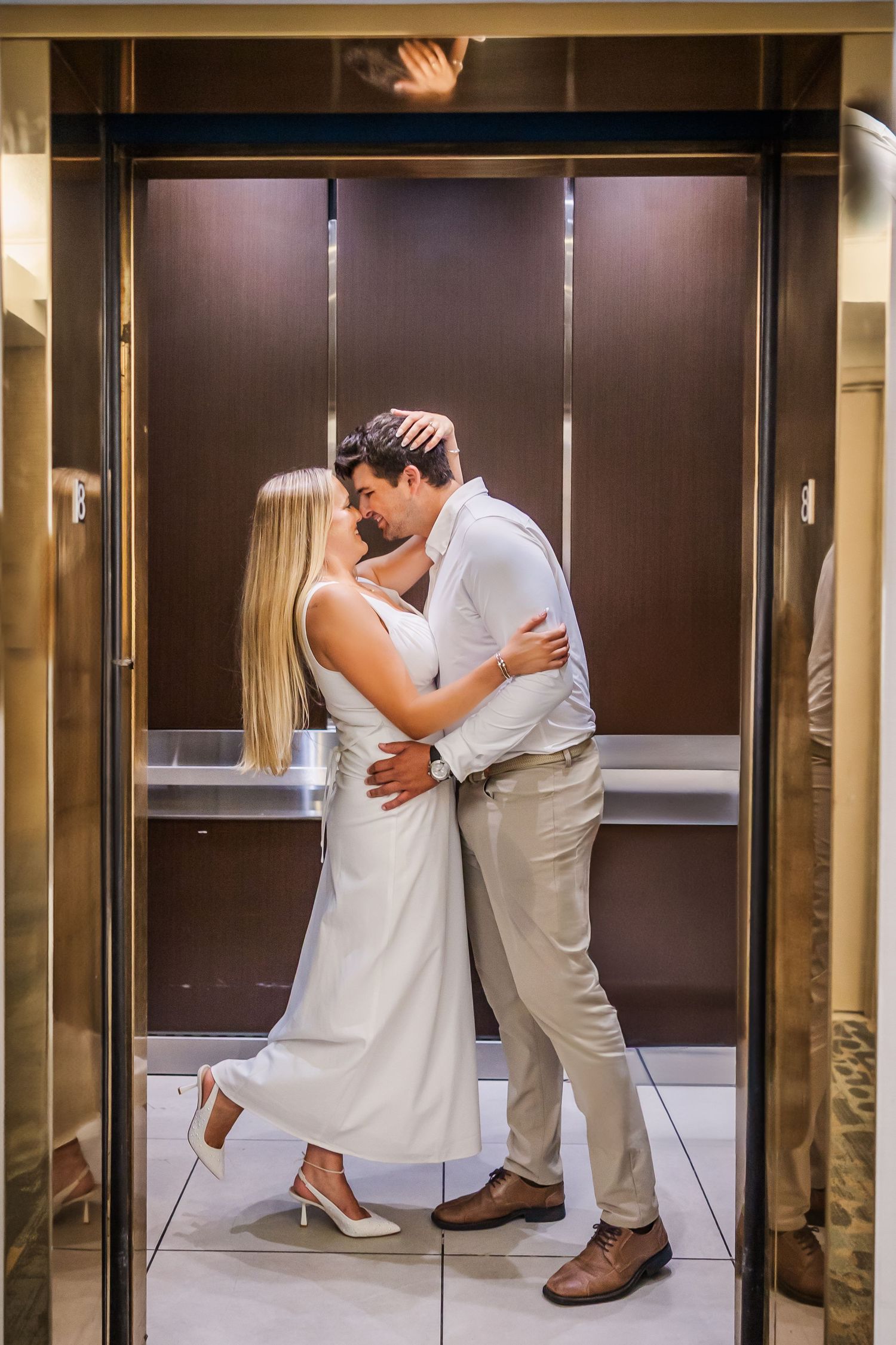A romantic couple embracing in an elevator, wearing white and beige formal attire.