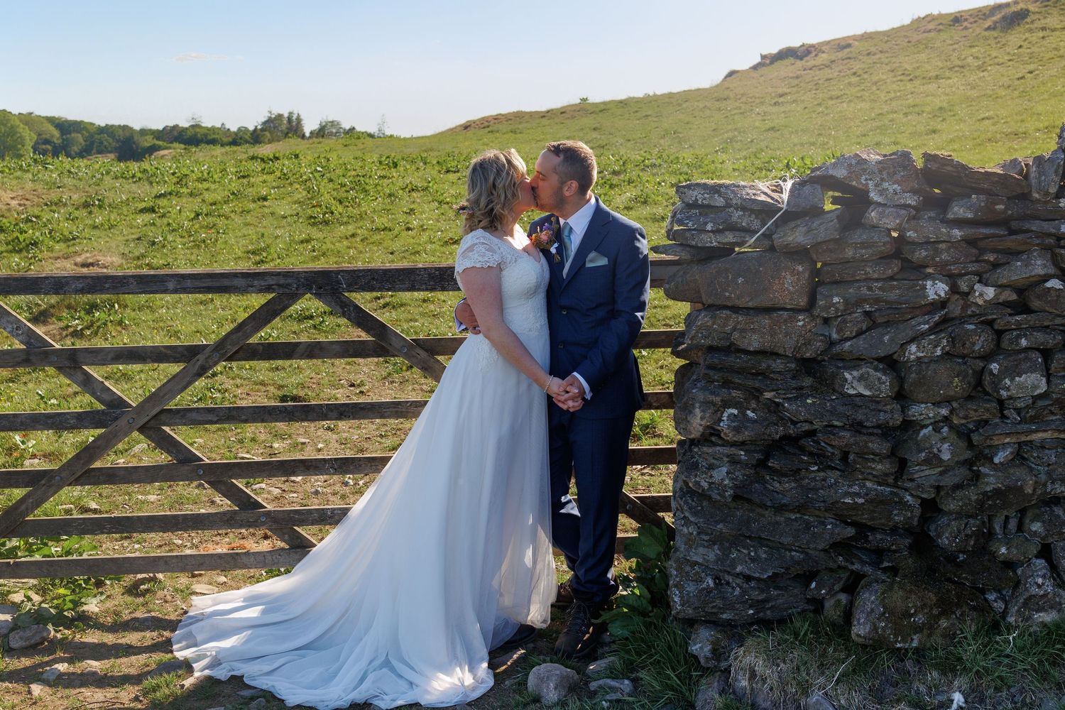 A romantic wedding couple embraces near a rustic stone wall and wooden gate in the countryside.