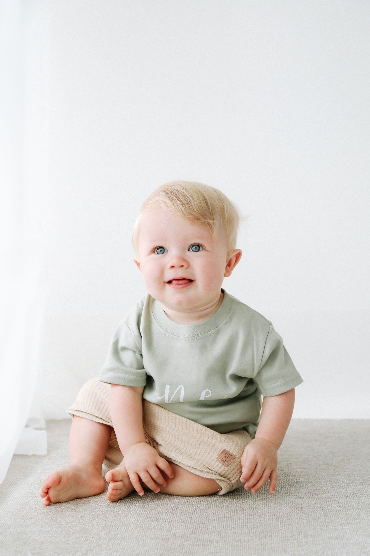 Happy baby wearing sage green t-shirt and beige shorts sits on gray surface against white background.