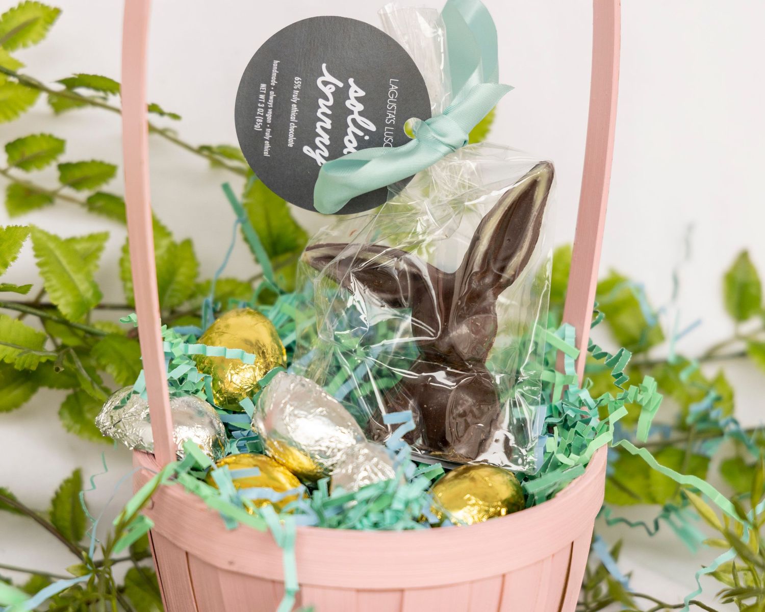 A pink Easter basket filled with green paper grass, chocolate eggs and bunny decorations sits among spring foliage.