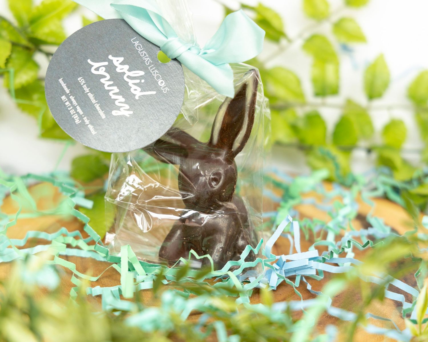 A chocolate Easter bunny wrapped in cellophane with a blue gift tag sits among spring decorations and greenery.