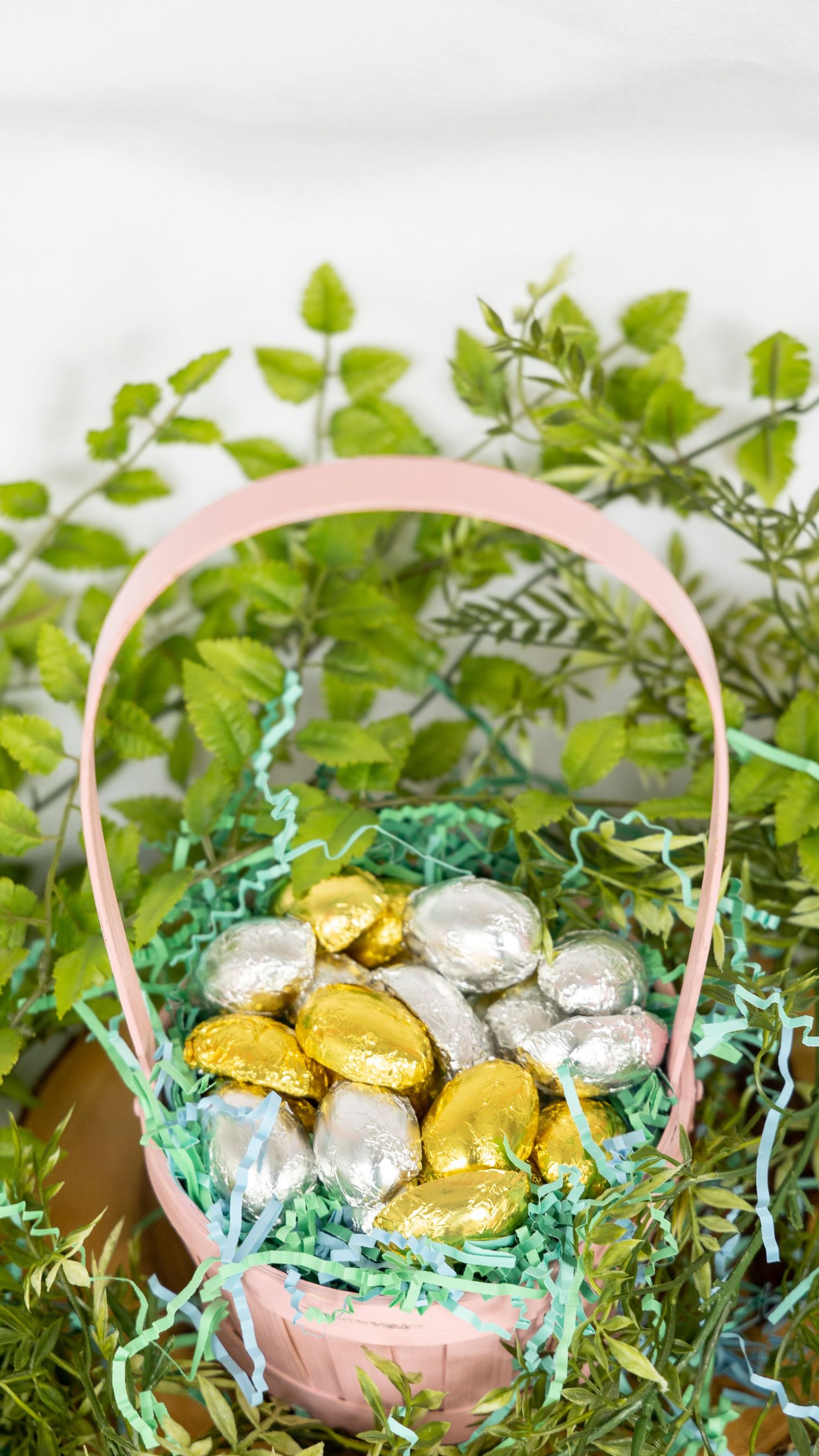 Pink Easter basket filled with gold and silver foil wrapped chocolate eggs nestled in green paper grass and ivy leaves.