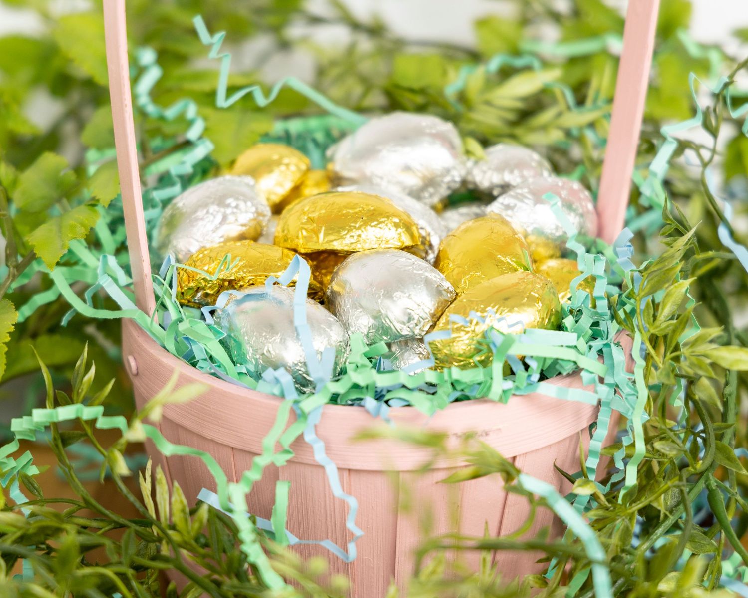 A pink Easter basket filled with gold and silver foil wrapped chocolates nestled in turquoise paper grass.