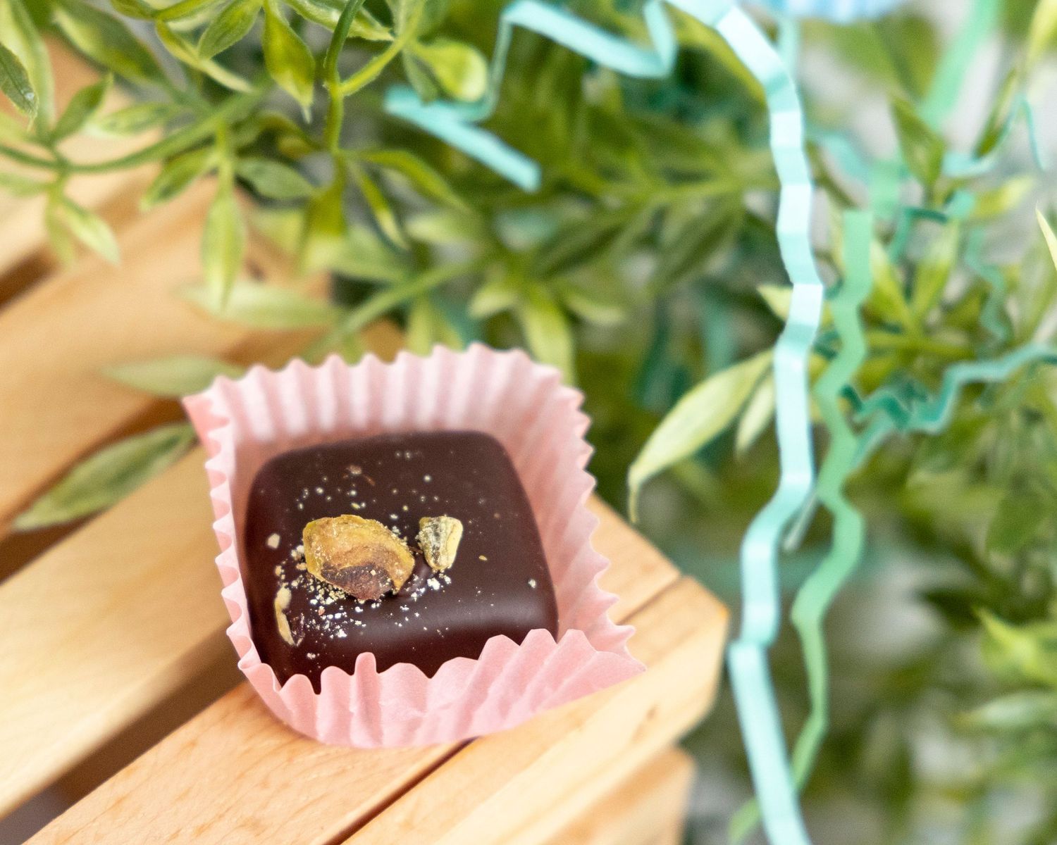 A dark chocolate truffle in a pink paper wrapper sits on a wooden surface with green foliage in the background.