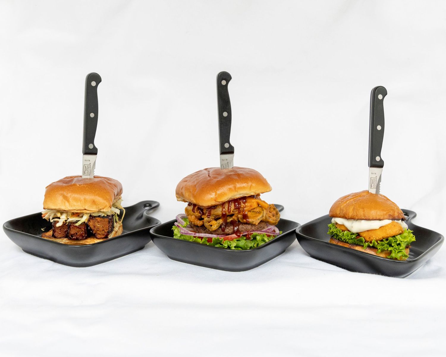 Row of gourmet hamburgers with toppings on black serving platters arranged in a progression from empty plate to finished burger.