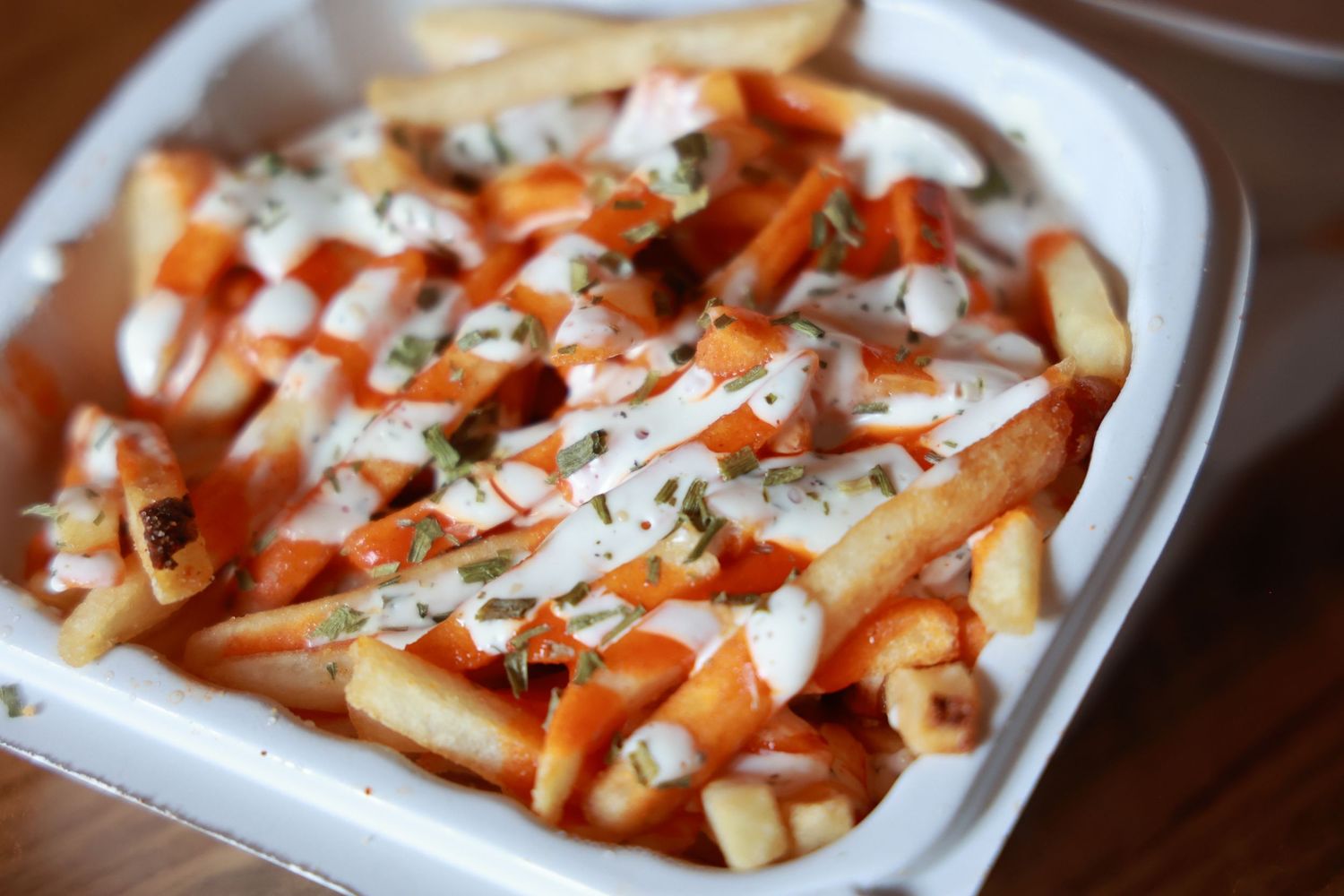 French fries topped with buffalo sauce, ranch dressing, and chopped herbs served in a white takeout container.