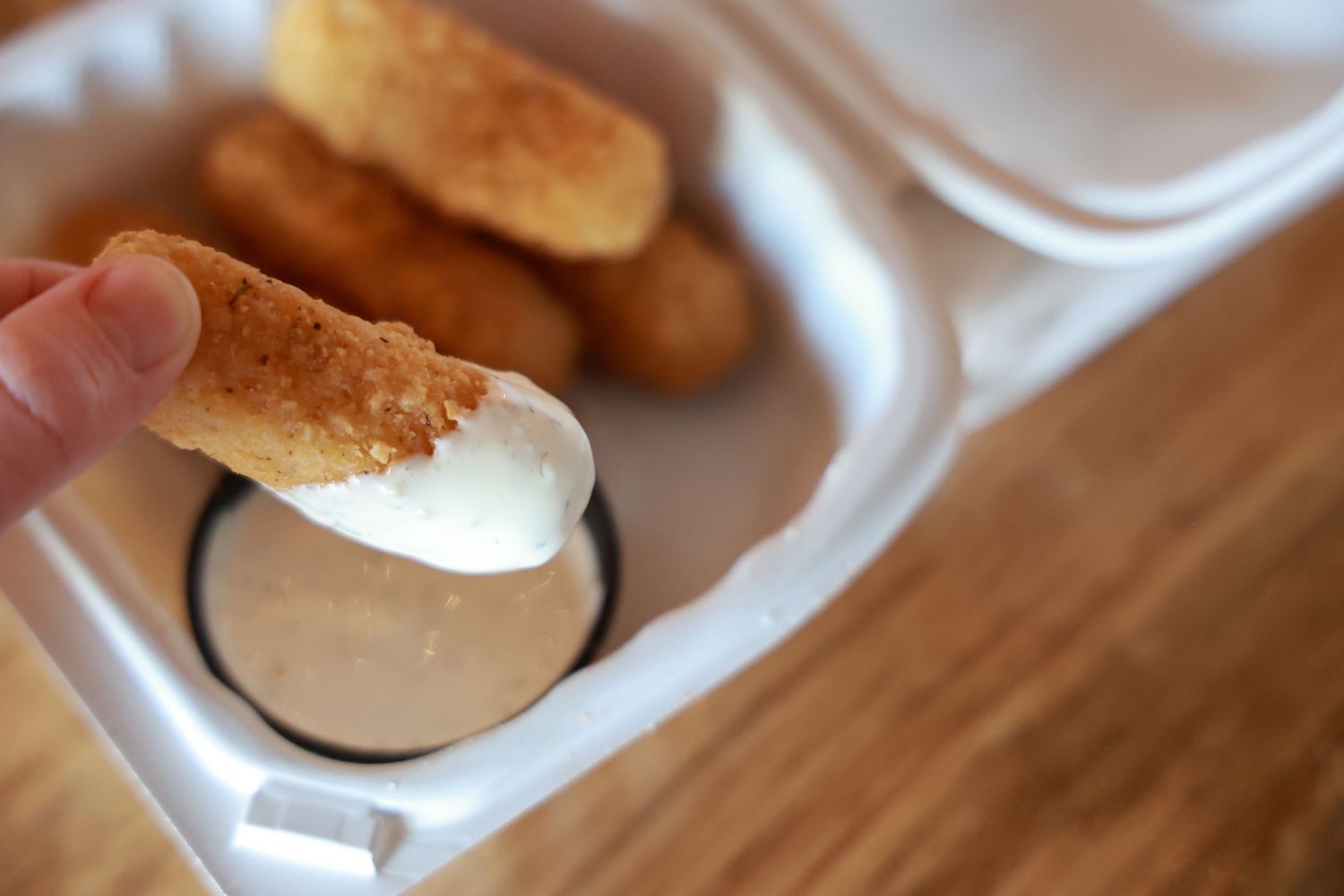 A hand dipping a crispy chicken tender into creamy sauce in a white takeout container.