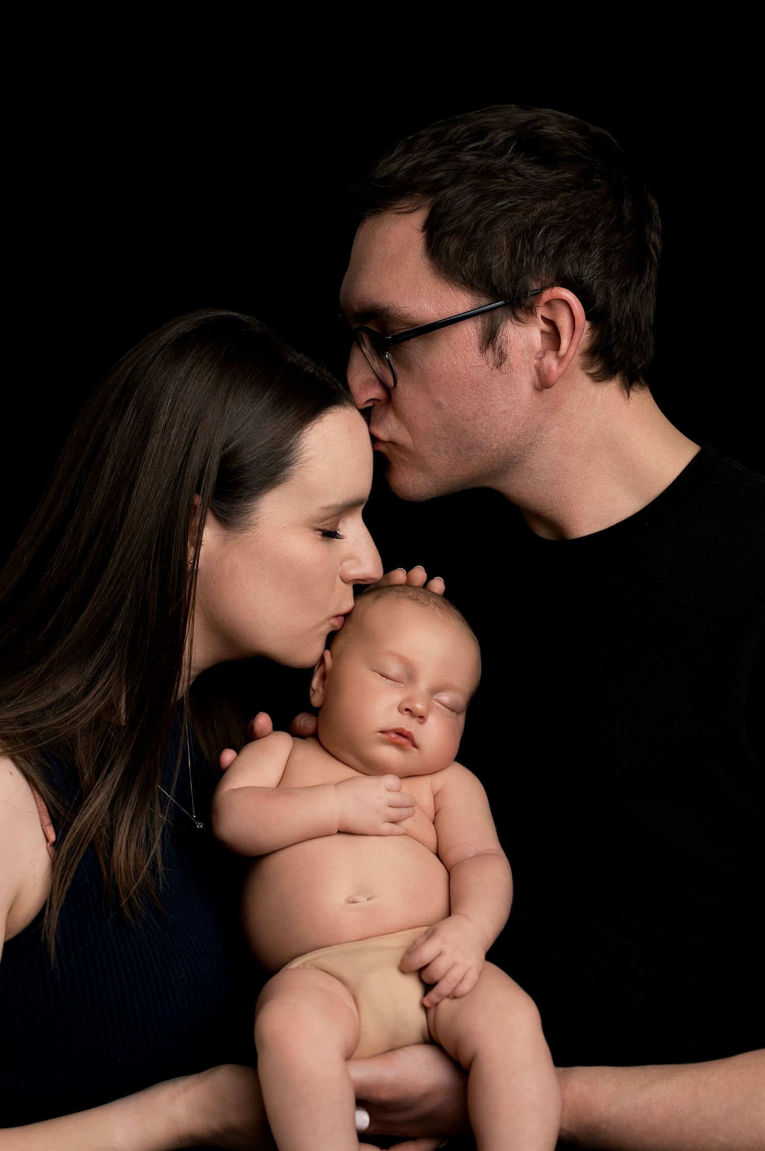 Intimate family portrait against dark background shows tender moment with sleeping newborn baby.