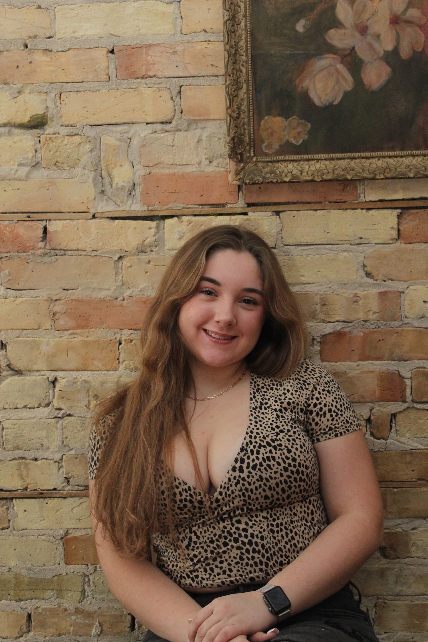 A woman wearing a leopard print top sits against a rustic brick wall with a floral painting visible in the background.