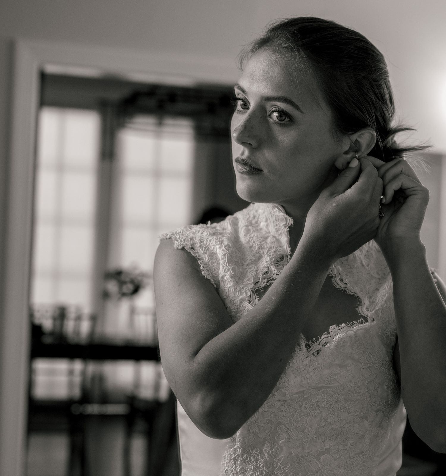 A bride in a white lace dress adjusts her formal wedding shoe while sitting in a room with decorative lamps and hardwood floors.
