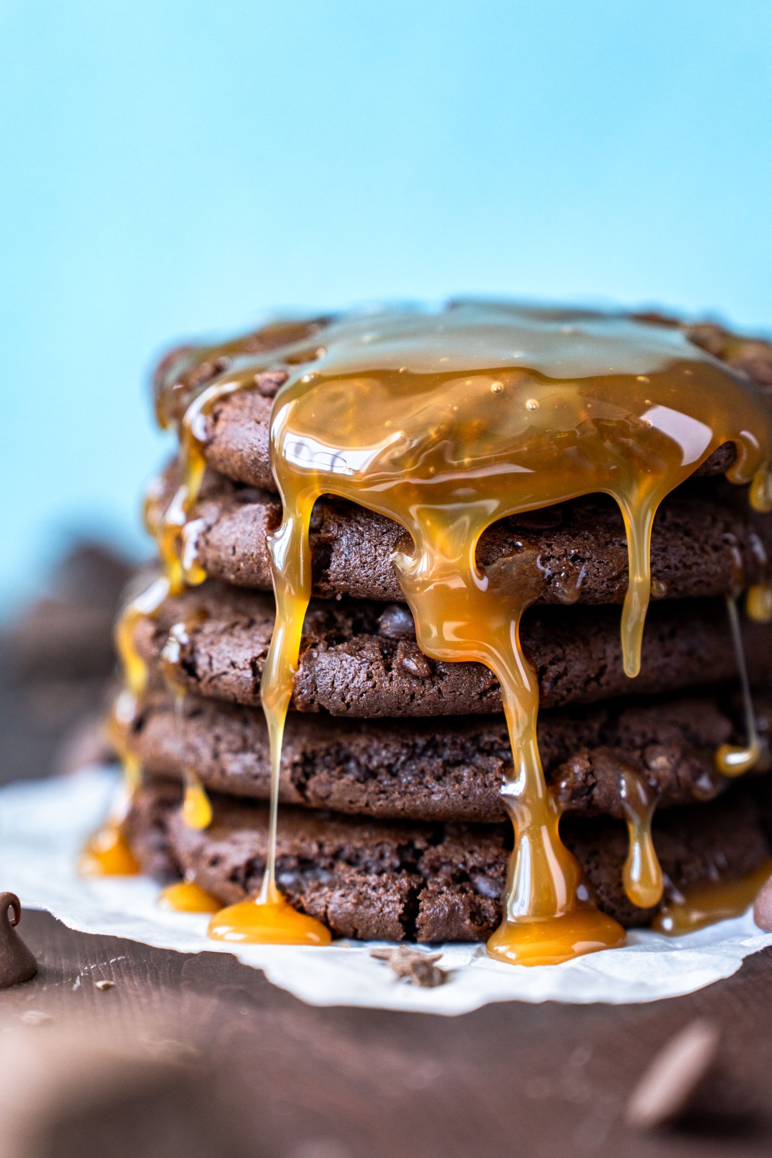 Chocolate cookies stacked on top of each other with caramel sauce dripping down the sides in front of blue backdrop.