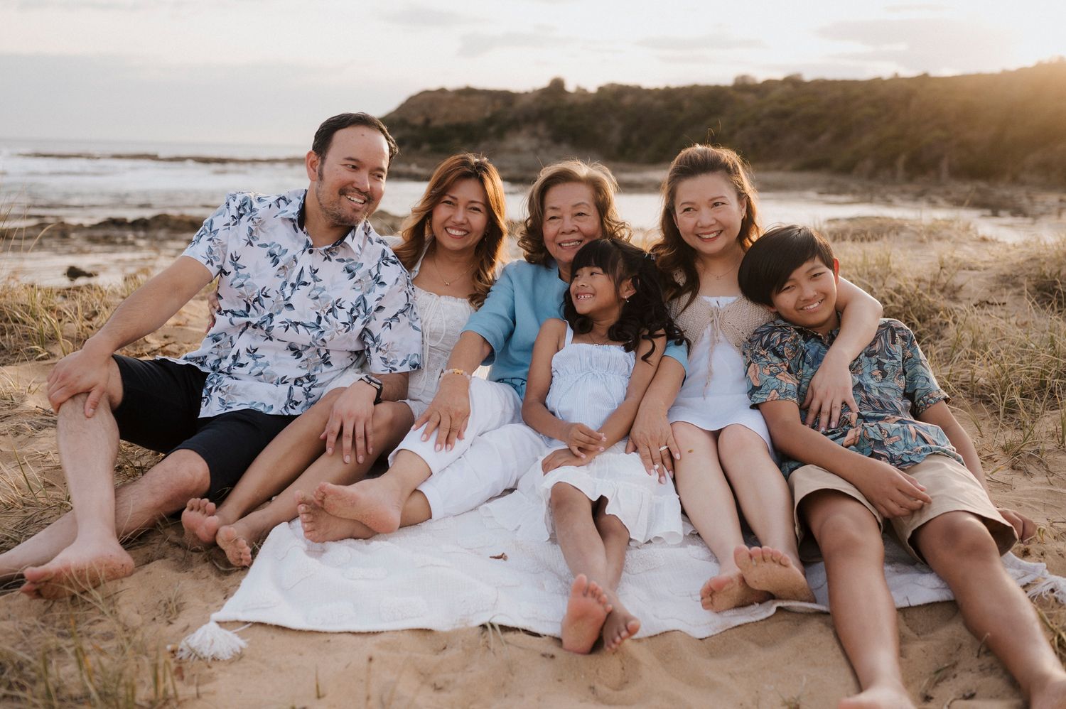 Four generations gather at sunset on a rocky beach, standing together barefoot with a warm golden glow around them.