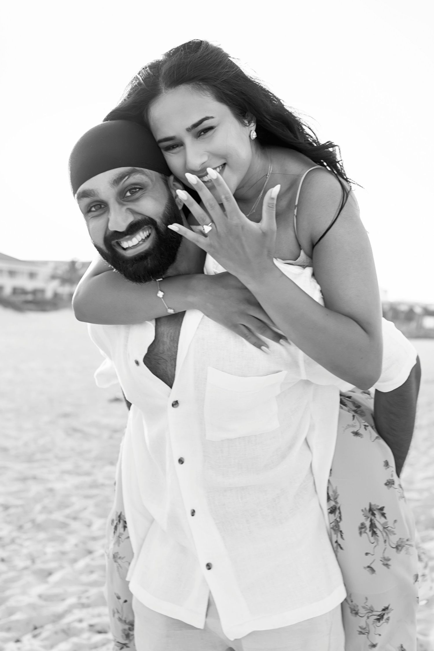 Black and white photo of a happy couple on a beach, woman showing engagement ring while getting a piggyback ride.