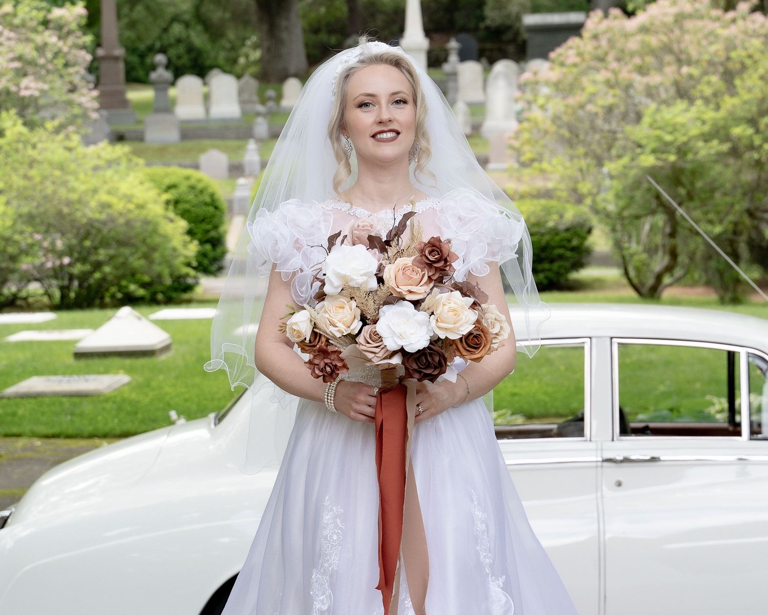 A wedding bouquet with blush pink and burgundy flowers is held against a white dress in an outdoor garden setting.