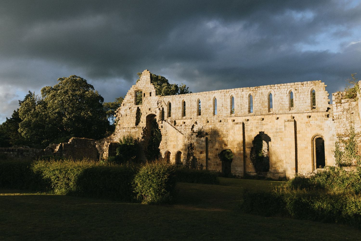 Ancient stone ruins stand against a dramatic stormy sky with sunlit walls stretching across a green landscape.