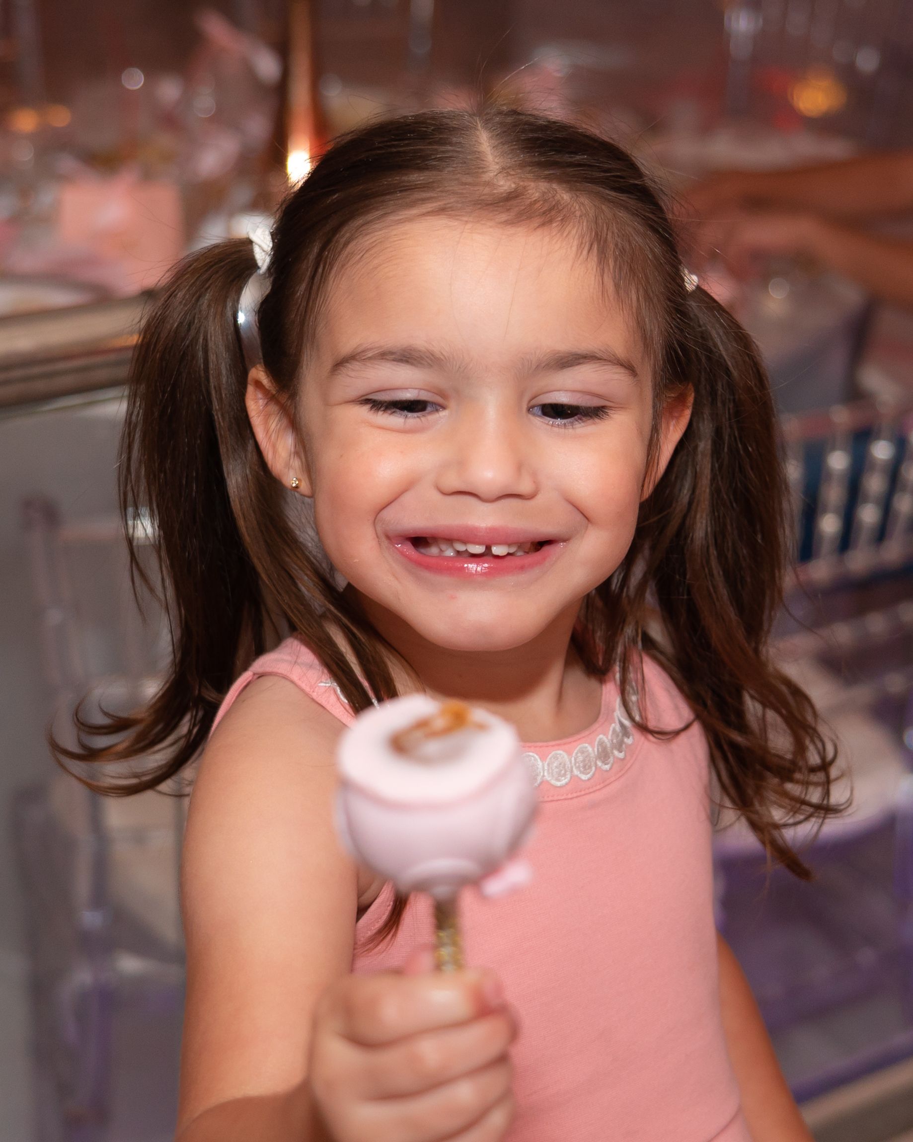 Adorable young girl with pigtails smiling and holding a pink cake pop at a celebration event.