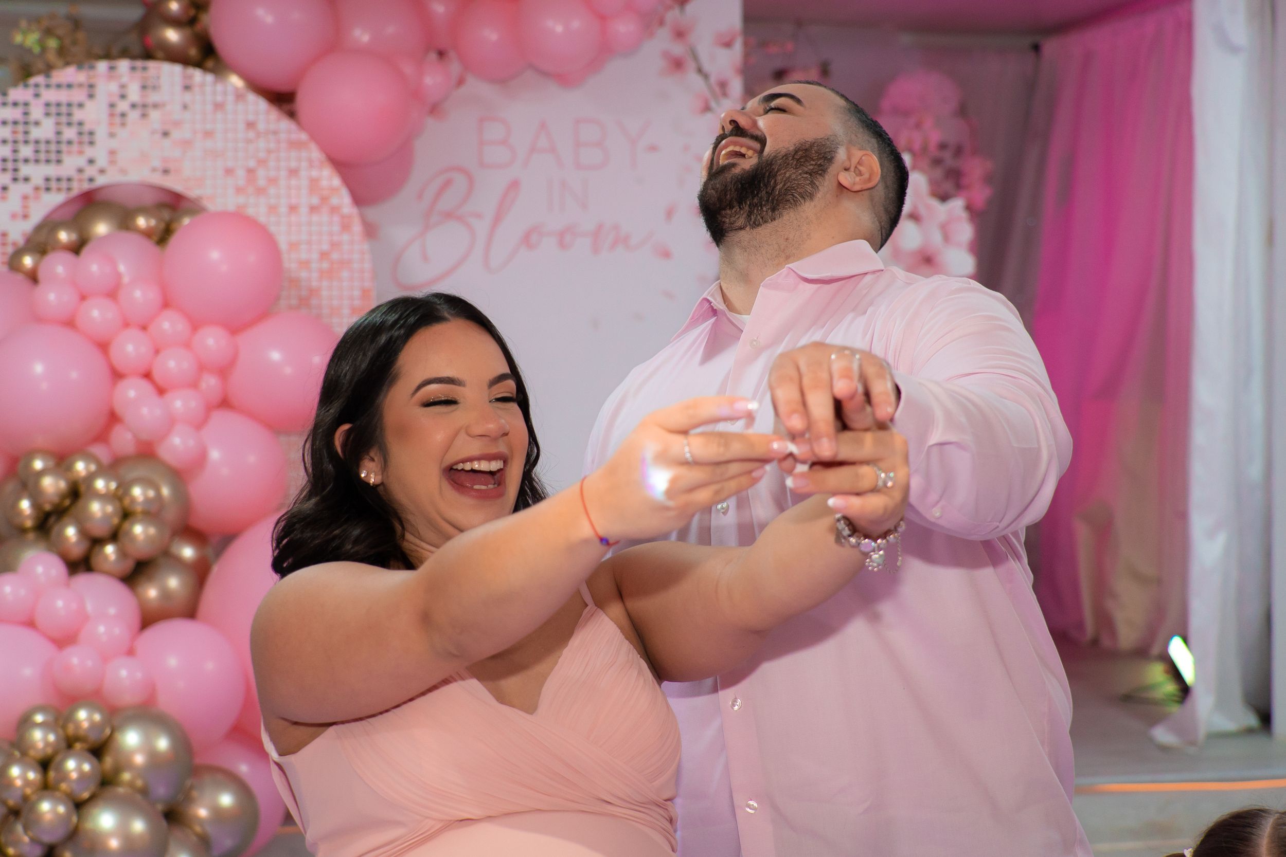 Couple laughing and celebrating together at a pink and gold baby shower with balloon backdrop.