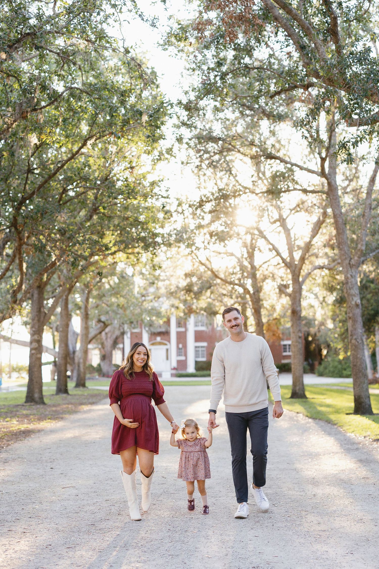 Family walking together on a winding sandy path through a tranquil park with sunlight filtering through oak trees.
